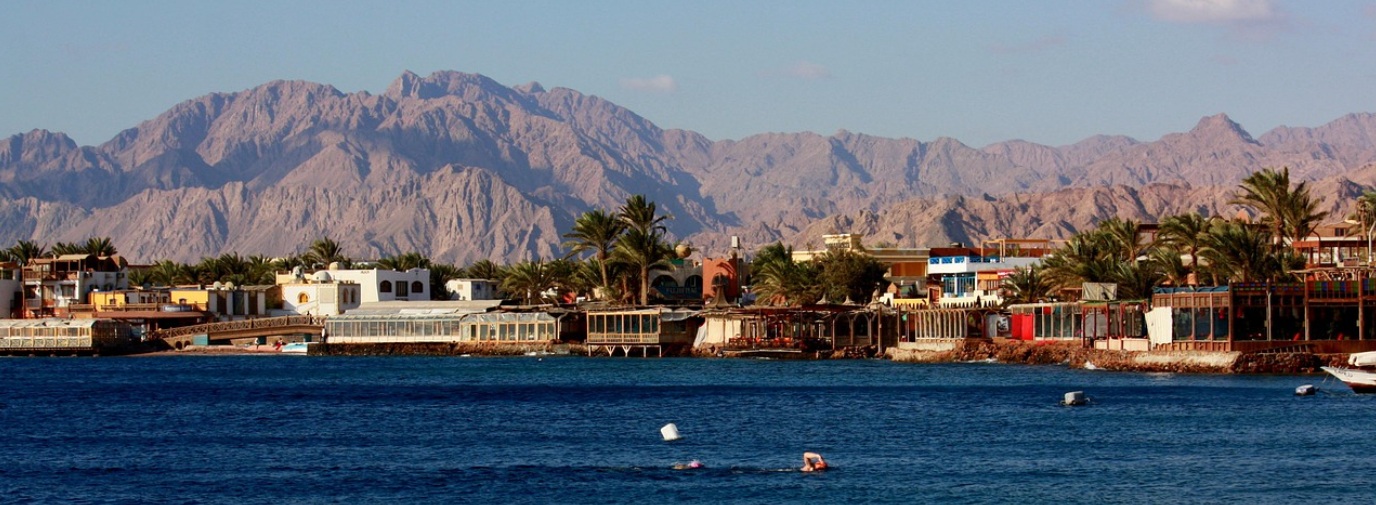 이집트 다합(Dahab) 라이트하우스 해변 전경. 홍해의 푸른 바다에서 수영하는 사람들과 해안가를 따라 늘어선 저층 호텔 및 레스토랑들, 그 뒤로 병풍처럼 펼쳐진 시나이반도의 웅장하고 거친 황금빛 산맥.