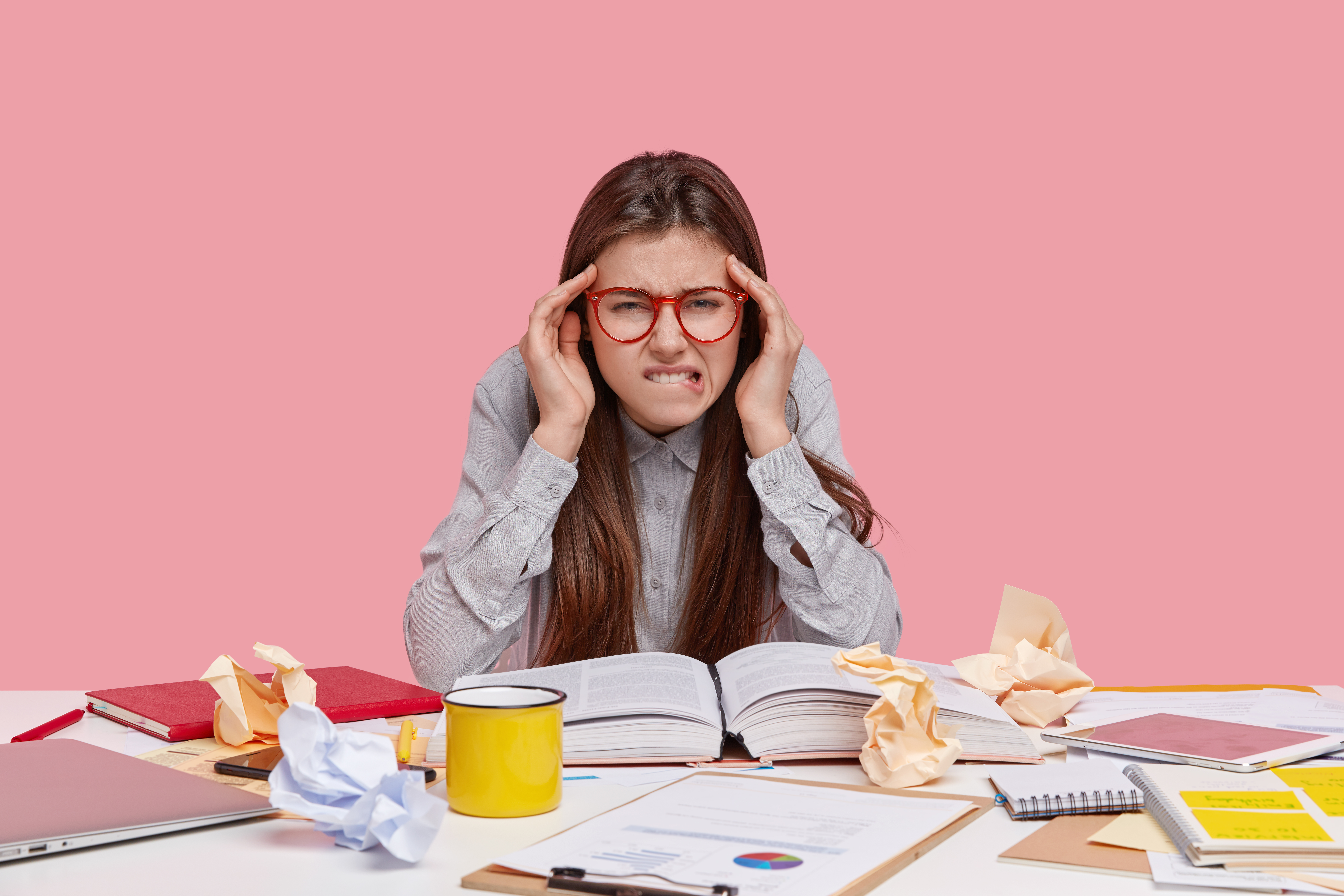 overworked-pretty-woman-bites-lower-lip-suffers-from-headache-reads-scientific-article-book-has-mess-table-1