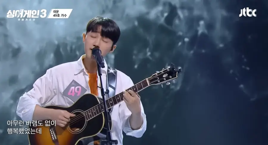 싱어게인3- 흰색셔츠안 주황색 티를 입고 눈을 감은 상태에서 스탠딩 마이크 앞에서 갈색기타을 치며 노래하는 49호