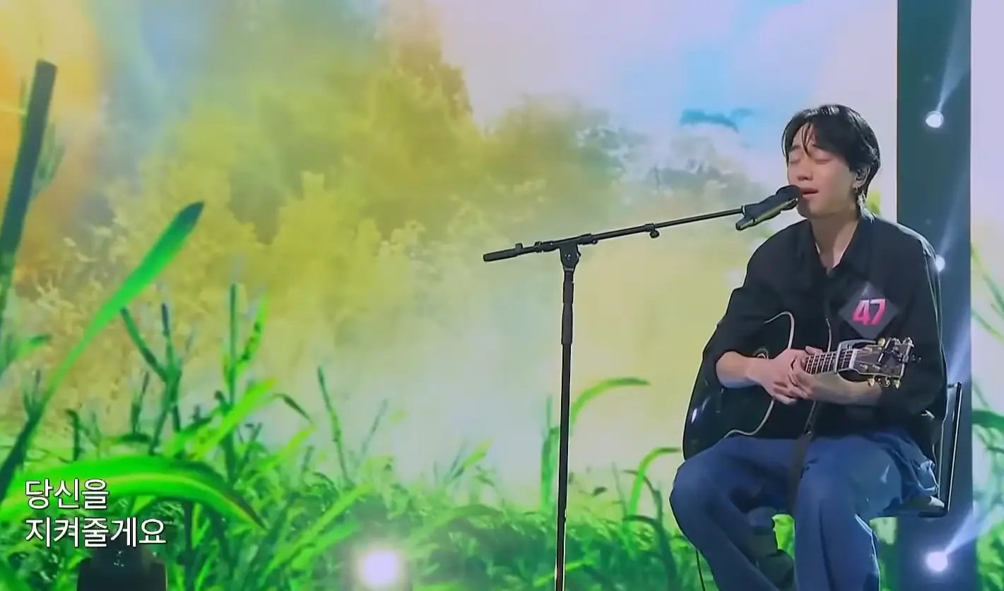 싱어게인3- 검은색 셔츠와 청바지에 가슴에 47번호표를 달고 검은색 기타를 감싸안고 스탠딩마이크를 앞에 두고 앉아 노래하는 47호 옆모습