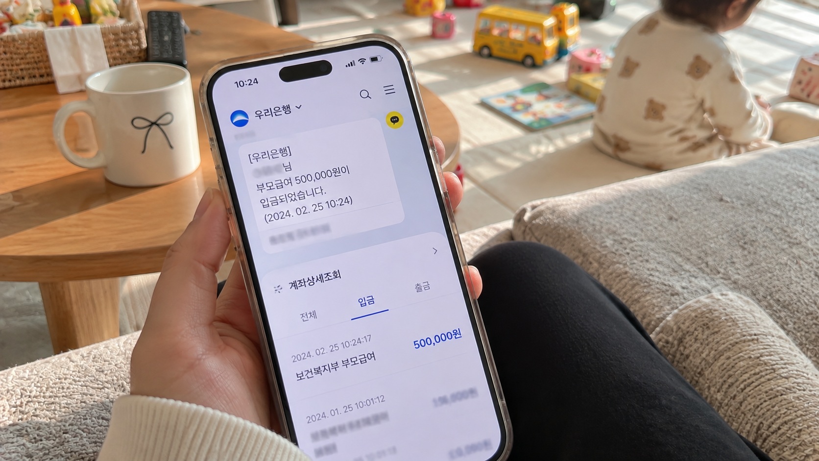 거실에서 스마트폰 은행 앱을 통해 부모급여 50만원 입금 알림과 계좌 거래내역을 확인하는 모습