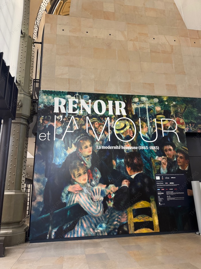 A large interior exhibition wall at Musee d'Orsay featuring Pierre-Auguste Renoir's painting and bold typography reading Renoir et l'amour