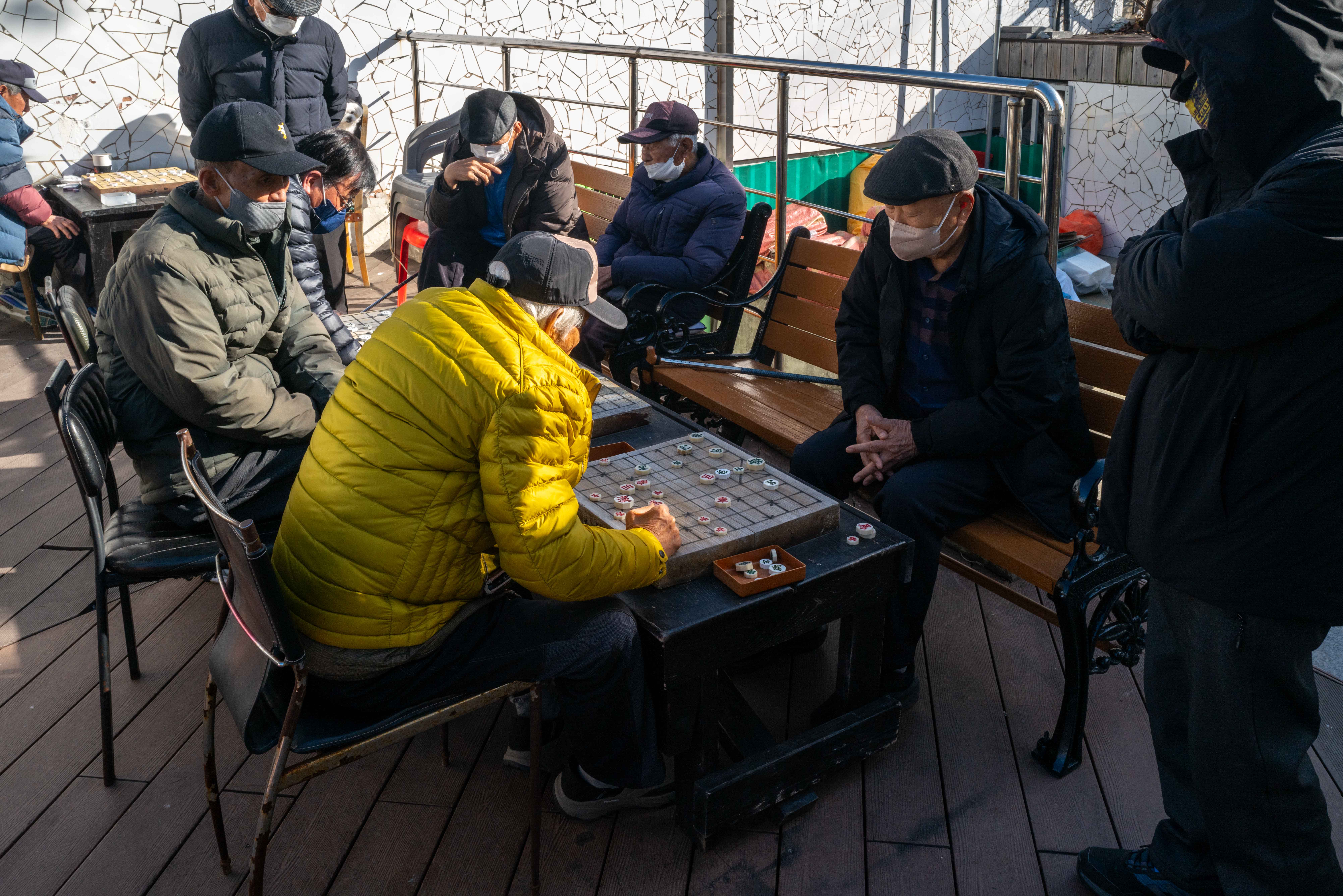 나란이 있는 3개의 테이블에 할아버지들이 앉아 장기를 두고 있다. 그리고 그 옆에는 그걸 구경하는 사람들이 서있다.