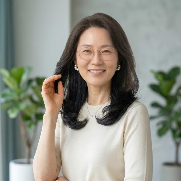 아이보리색 상의를 입고 안경과 진주 귀걸이를 착용한 채 밝고 자신감 있게 미소 짓고 있는 중년 여성의 인물 사진