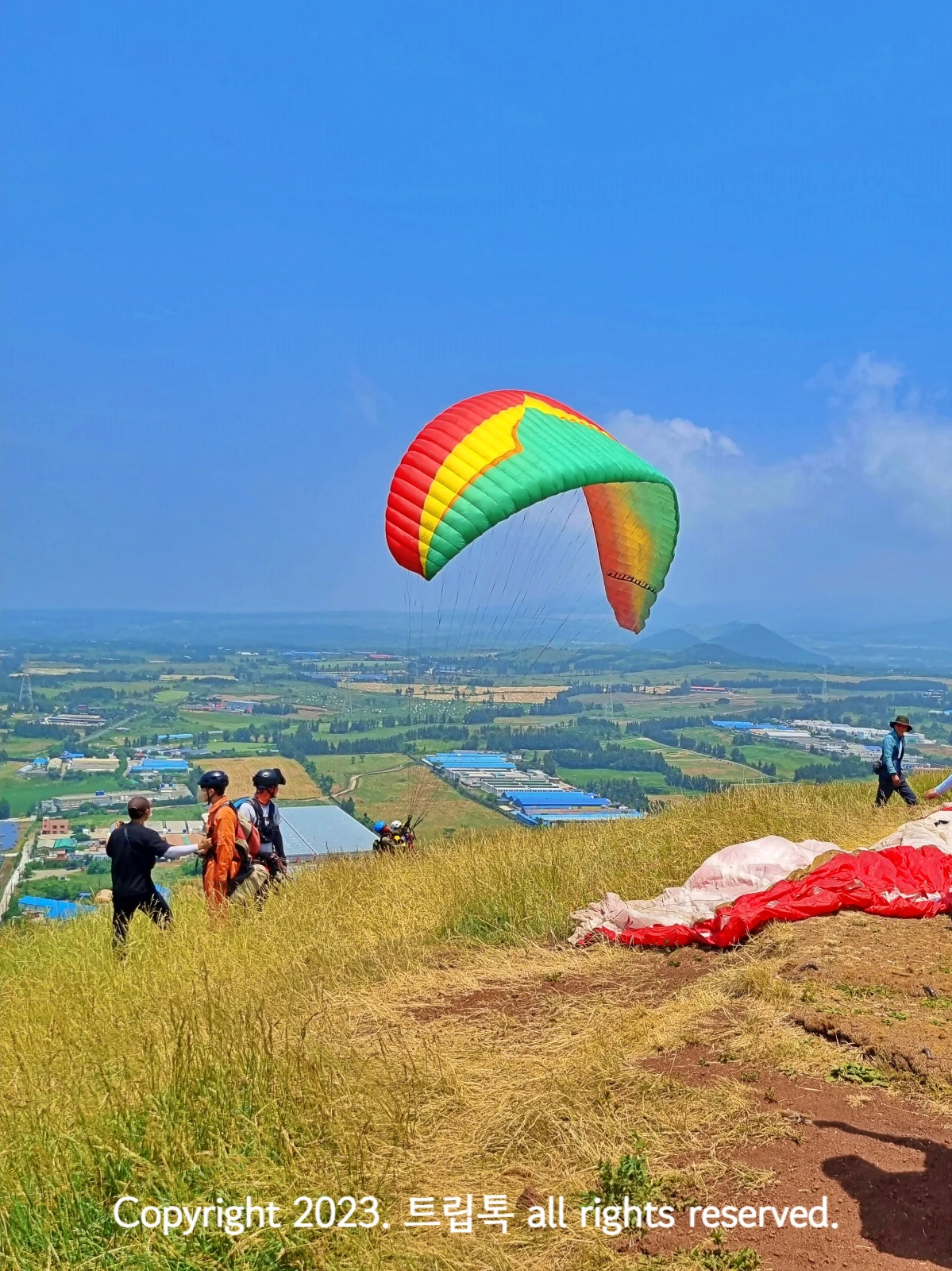 파란 하늘에 빨강,노랑,초록색 무늬 패러글라이딩이 펼쳐져있고 땅에는 빨간게 널어져있다.