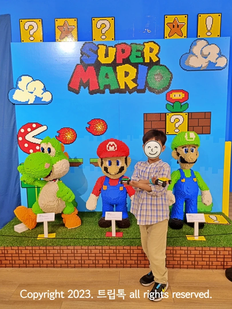 브릭으로 만든 연두생 공룡과 빨간색 마리오와 연두색 루이지와 그 앞의 한 아이가 있다