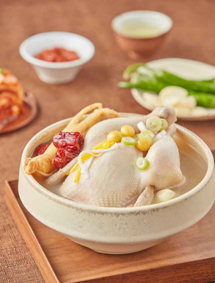 A steaming bowl of Samgyetang (Korean ginseng chicken soup) with jujube, ginkgo nuts, green onion, and ginseng root. Classic Korean side dishes like kimchi, green chili peppers, and garlic are seen in the background.