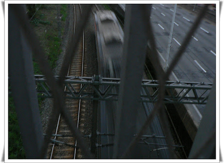 철조망 틈 사이로 촬영된 이미지로, 중앙에는 어두운 철도 선로 위를 빠르게 지나가는 기차의 흐릿한 형체가 보인다. 오른쪽에는 한산한 자동차 도로(강변북로)가 병행하고 있고, 위에는 철제 구조물이 가로지르고 있다. 전체적으로 어둡고 긴박한 느낌의 장면이다.