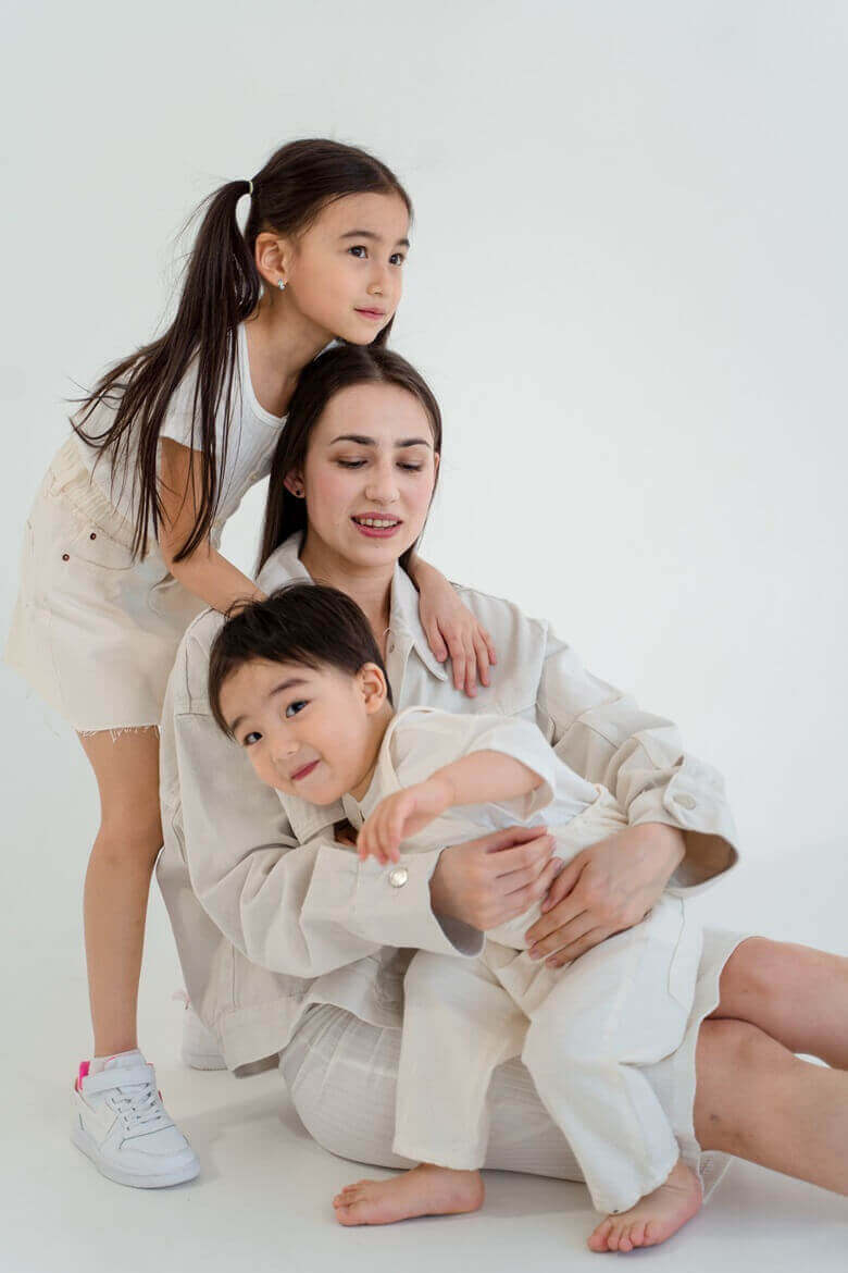 엄마가 방 바닥에 앉아있고 3살정도로 보이는 아들이 엄마 무릎에 앉아서 엄마품에 안겨있고 5살정도로 보이는 딸이 엄마 등뒤에서 껴안고 있는 모습