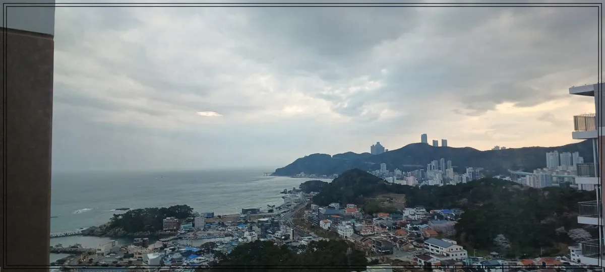 먼곳에-바다와-낮은-산-가깝게-마을이-보임-군데군데-낮은-구릉이-있음-하늘에-구름이-잔뜩-있지만-구름사이로-빛이-보임