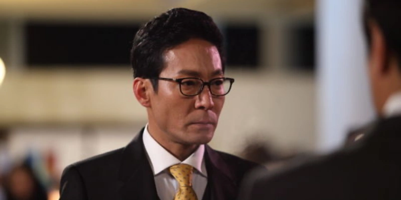 어느 실내를 배경으로 머리를 위로 올리고 안경과 검은 정장을 입고 서 있는 드라마 7인의 탈출의 구강재 캐릭터