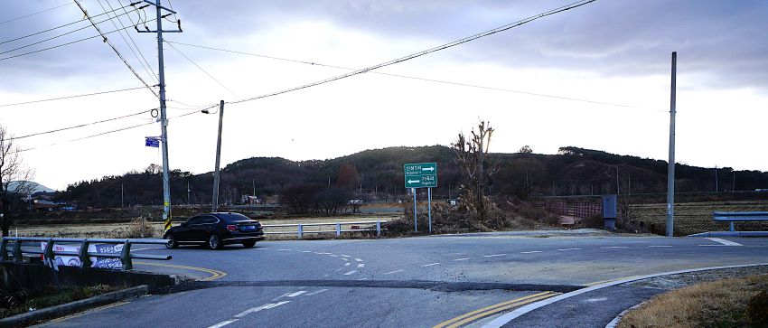 삼거리, 가운데에 녹색바탕에 흰 화살표 표시된 교툥 안내판, 왼쪽으로 기곡리로 가는 방향, 가운데 검은 승용차 1대 좌로 진행 중, 뒤로는 야산, 하얀 하늘,