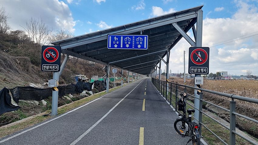 옛 선로위에 깔끔하게 포장된 자전거도로, 이어지는 지붕형 태양광발전 패널판, 좌 우에 들판, 파란 하늘에 흰구름, 왼쪽 보도는 자전거 통행 행금지, 우측 자전거길은 보행금지,