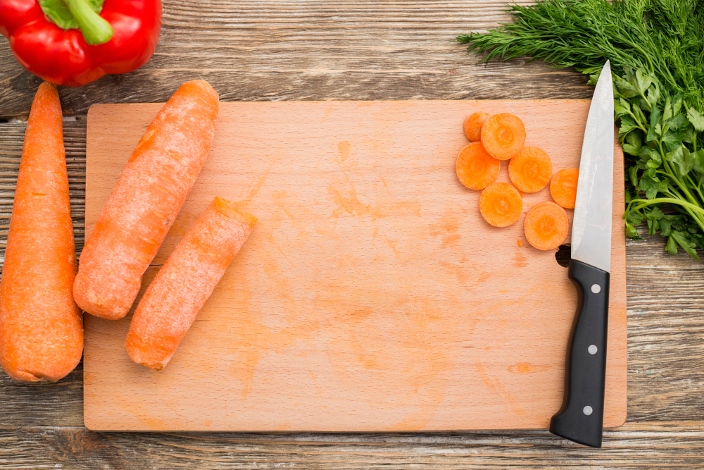 나무 도마 위에 썰린 당근과 칼이 놓여 있고, 도마에는 당근의 주황색 얼룩이 남아있다. 주변에는 빨간 파프리카와 신선한 허브가 배치되어 있는 주방 조리 장면.