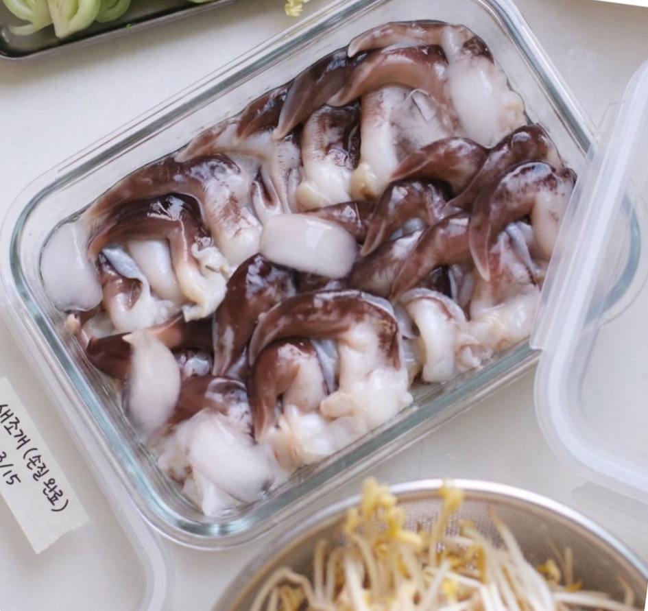 하얀 조리대 위, 투명한 유리 밀폐용기에 가득 담긴 손질된 새조개 사진. 용기 옆에는 '새조개 (손질 완료) 3/15'라고 적힌 종이 테이프 라벨이 붙어 있고, 주변으로 신선한 청경채와 채반에 담긴 숙주나물이 함께 놓여 있는 정갈한 식재료 준비 모습.