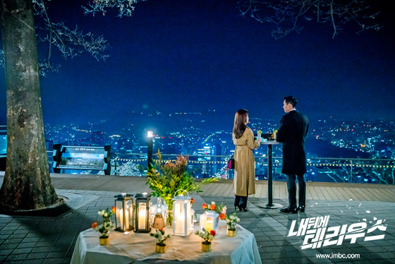 도시의 야경이 내려다보이는 야외 테이블 옆에 서 있는 두 인물