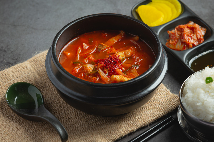 검은 바닥 위에 헝겊깔판을 깔고 그 위에 숟가락, 젓가락과 검고 둥그런 뚝배기에 김치찌개를 가득 담아놓고, 그 옆에 밥을 담은 쇠그릇과 김치와 반찬을 담은 그릇을 두고 찍은 사진