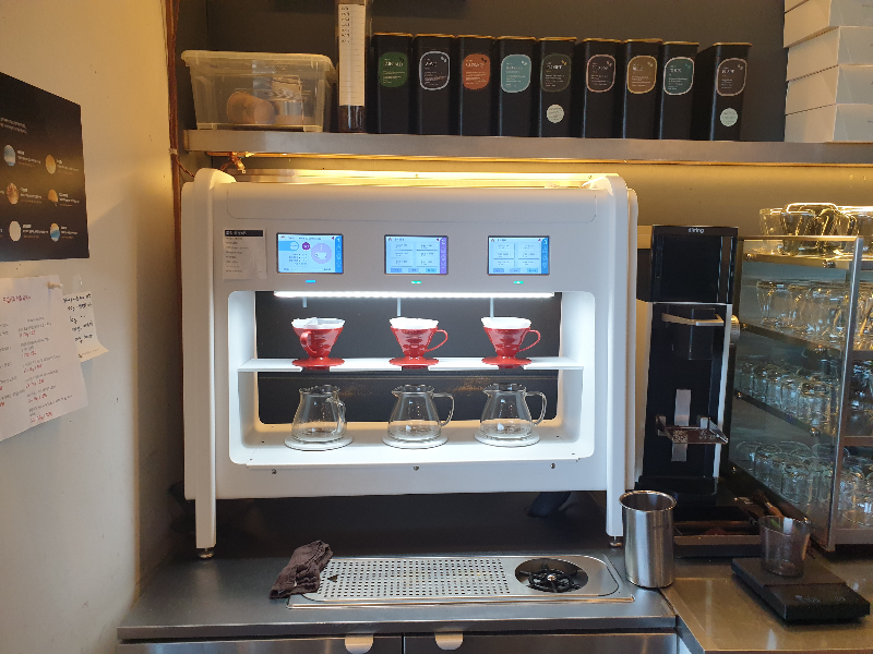 한 번에 세 잔을 추출할 수 있는 자동 브루잉 머신, 빨간 드리퍼와 서버 그리고 머신의 추출구와 조작 버튼이 보인다.
