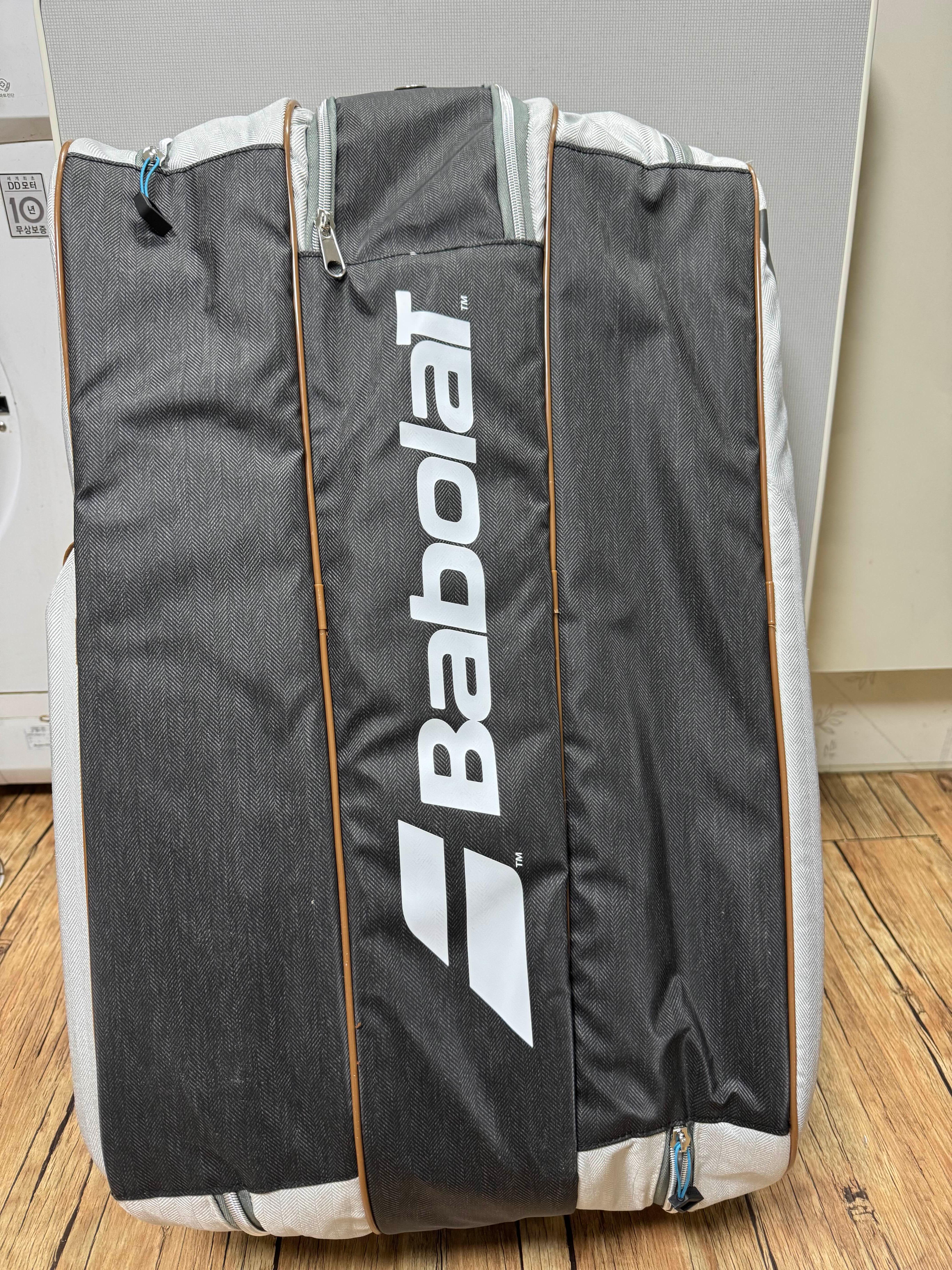 세로로 세워진 바볼랏 (babolat) 브랜드의 테니스 가방. 짙은 회색과 연회색 배색에 흰색 바볼랏 로고가 크게 새겨진 디자인.