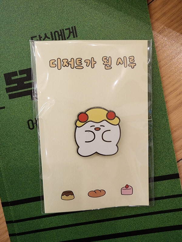 &#39;디저트가 된 시루&#39; 뱃지. 팔다리가 달린 찹쌀떡 같은 캐릭터가 눈을 감고 함박 웃음 짓고 있다. 머리에는 노란 소스가 뿌려져 있고 빨간 색 동그란 핀을 양 쪽에 달고 있음.