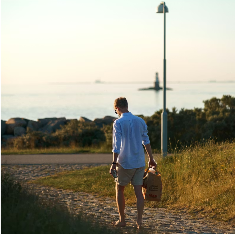 해질 무렵 해변길을 걷는 중년 남성의 뒷모습. 한 손에는 식료품 봉투를 들고 있으며, 편안한 복장으로 여유로운 산책을 즐기고 있다.