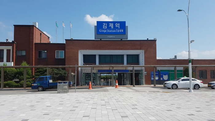 전북 김제역 전경. 조용하고 소박한 시골역 플랫폼에서 여행자가 걷고 있는 모습. 하늘은 맑고 배경에는 들판과 마을 풍경이 펼쳐진다.”