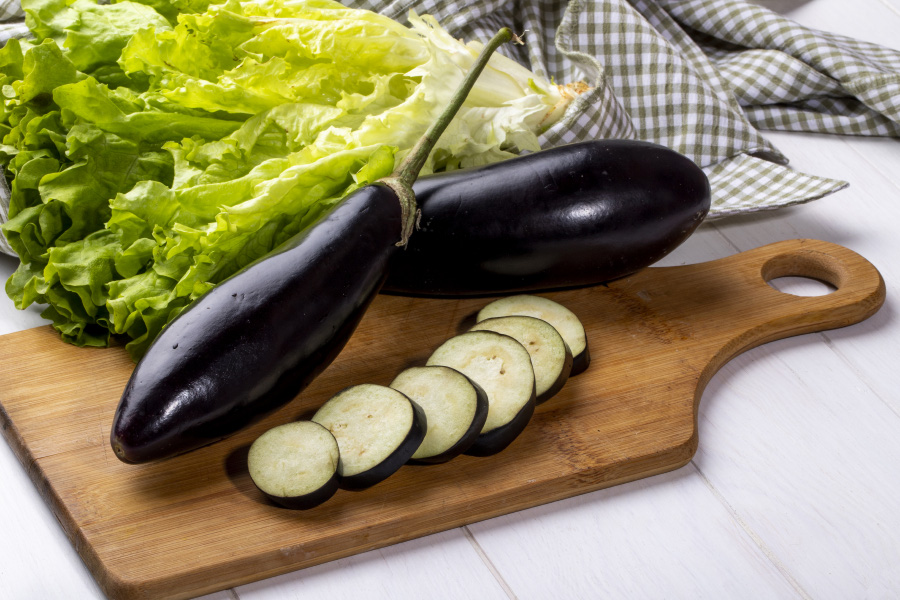 하얀 바닥 위에 나무 도마를 놓아두고 그 위에 가지 2개와 썰어 놓은 가지 조각 6개를 놓아두고, 옆에는 상추를 몇 개 놓아두고 찍은 사진