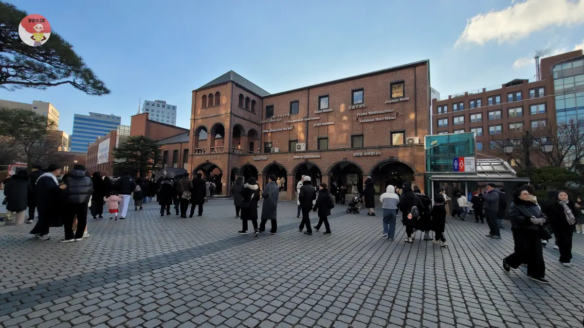 명동성당 앞 광장, 다양한 언어로 '메리 크리스마스'가 적힌 벽돌 건물과 겨울 옷차림의 사람들