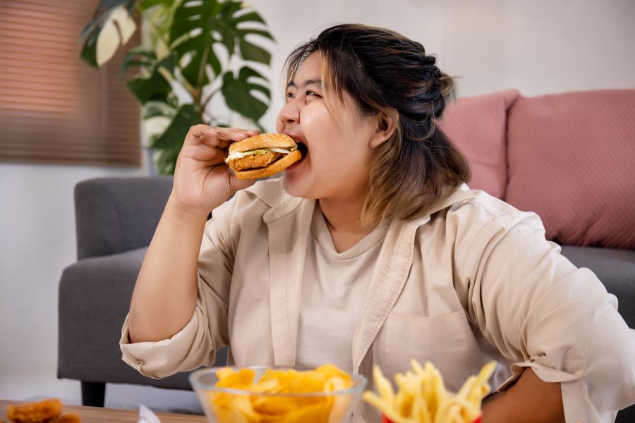 비만한 여성이 거실의 쇼파 앞에 앉아서 햄버거를 먹고 있고, 테이블 위에는 나초와 감자튀김 등을 놓아두고 있는 것을 찍은 사진