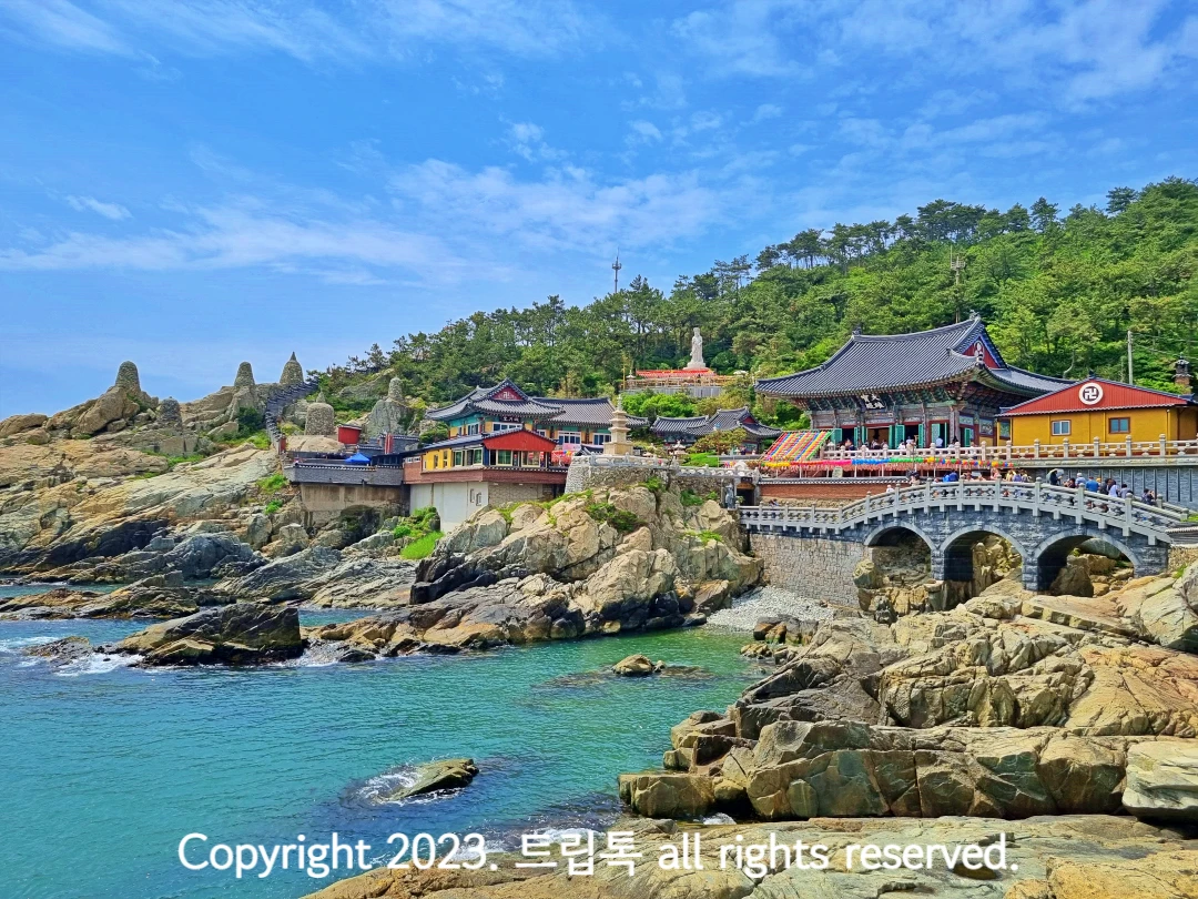 푸른 하늘아래 기와지붕의 절과 커다란 돌다리가 있고 그 아래 부산 푸른 바다가 있다.