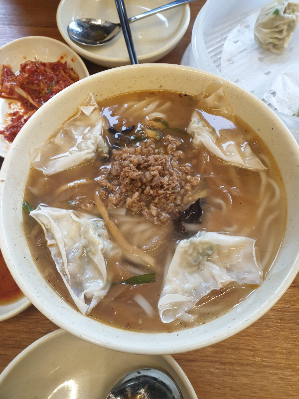 테이블 위에 칼국수. 칼국수 안에 물만두 네 개와 고기 고명이 보인다.