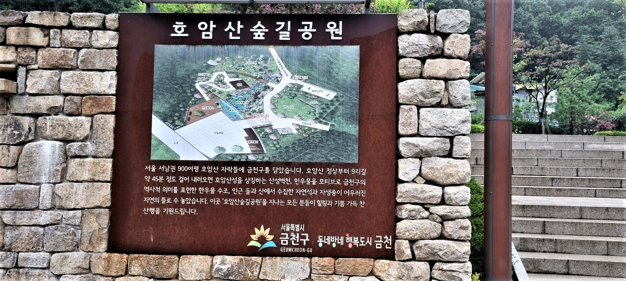 돌담에 부착된 호암산 숲길공원 안내판, 진한 갈색 판에 흰글씨, 우측은 공원에 오르는 계단,