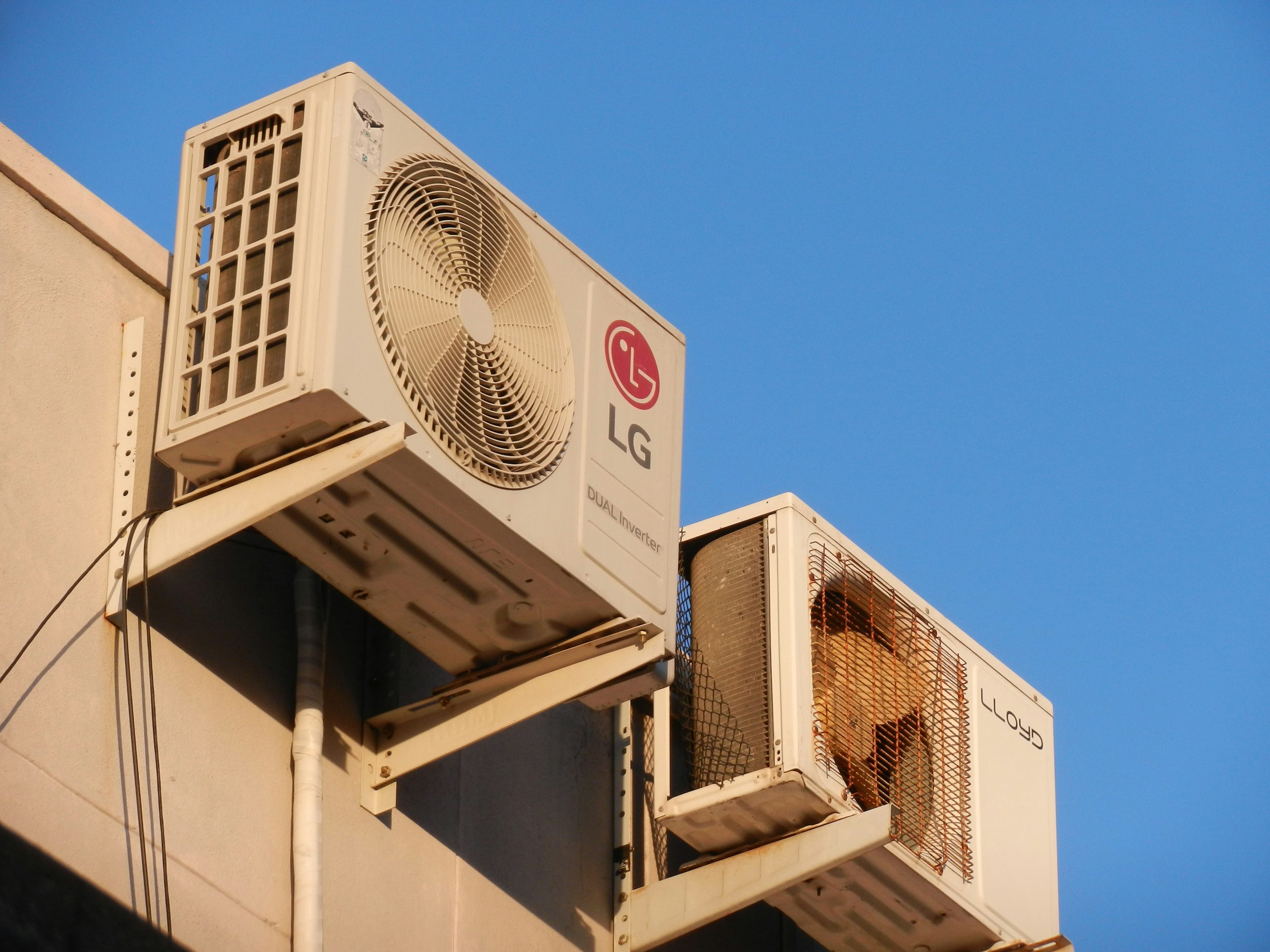 맑고 푸른 하늘을 배경으로 건물 외벽에 LG DUAL Inverter 에어컨 실외기와 Lloyd 실외기가 나란히 설치된 모습