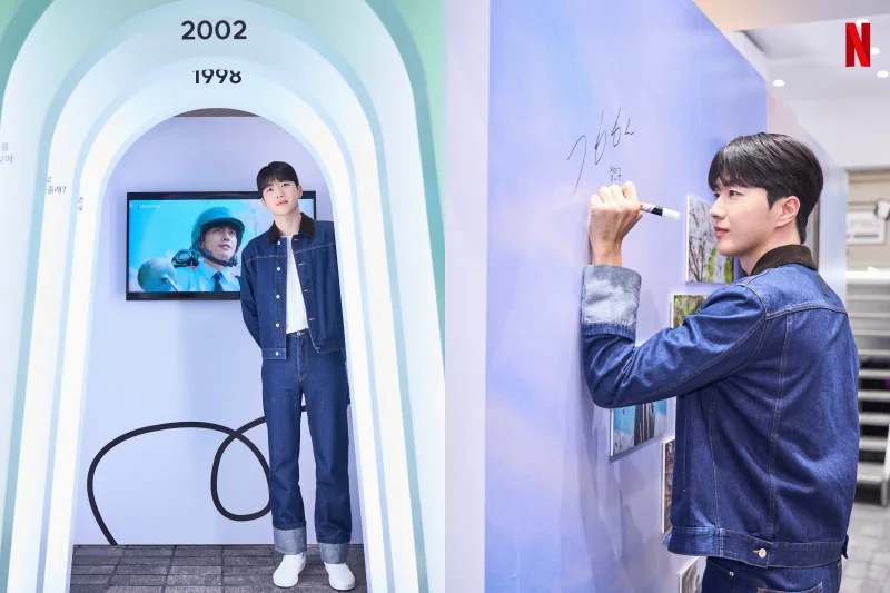 어느 실내를 배경으로 청바지와 청 자켓을 입고 벽면에 싸인을 하고 있는 배우 강훈