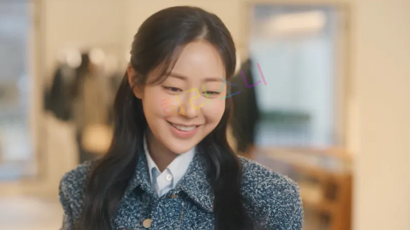실내에서 독고세리(신슬기)가 미소를 짓고 있다. 그녀는 파란색 트위드 재킷과 흰색 블라우스를 입고 있으며&amp;#44; 반묶음 스타일의 긴 머리를 하고 있다. 배경은 의류 매장처럼 보이며&amp;#44; 행거에 옷들이 걸려 있다. 