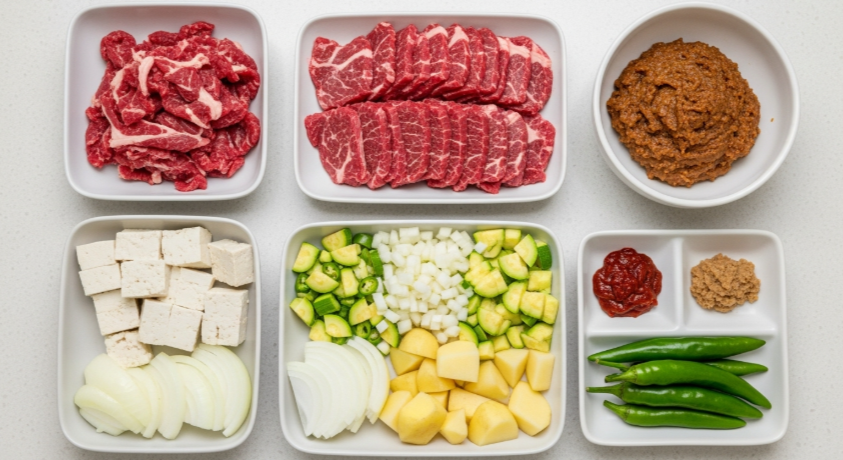 강된장에 사용될 차돌박이, 깍둑 썬 두부, 잘게 다진 양파, 감자, 호박, 고추 등 모든 재료와 된장 양념이 깔끔하게 준비된 평면 사진