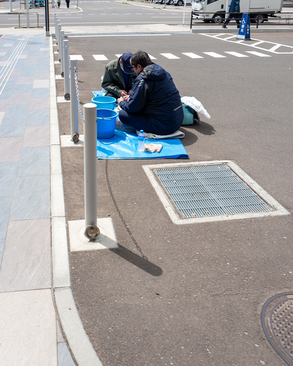 아스팔트 위에서 두 명의 남자가 바닥에 앉아 어떤 작업을 하고 있다. 28mm 화각으로 다소 멀리서 찍어 그들이 무엇을 하고 있는지까지 도드라지게 보이지는 않는다.