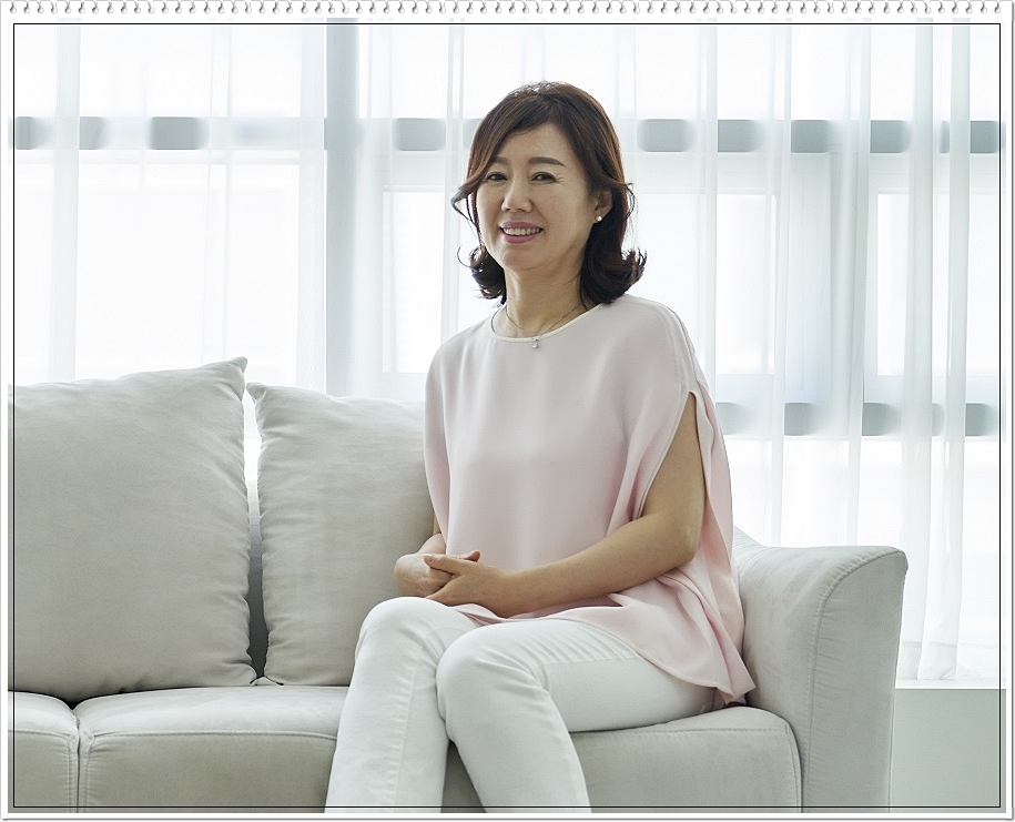 소파에 앉아 정면을 바라보며 미소를 짓고 있는 중년 여성의 모습