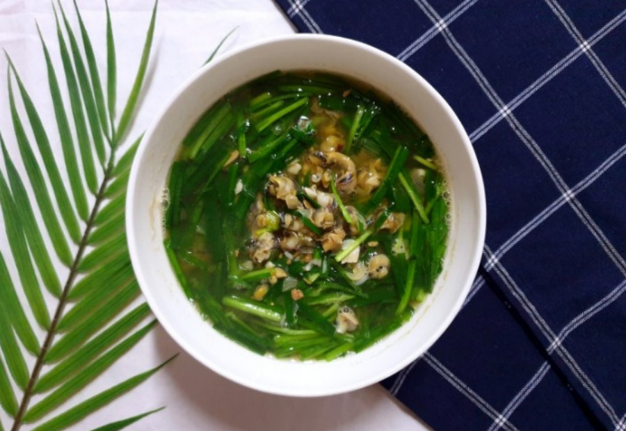 하얀 바닥에 왼쪽에는 나뭇잎을 오른쪽에는 체크무늬 깔개를 깔고 가운데에 동그랗고 하얀 그릇을 놓고, 거기에 부추와 올갱이 그리고 된장을 풀어서 만든 된장국을 가득 담아두고 위에서 찍은 사진