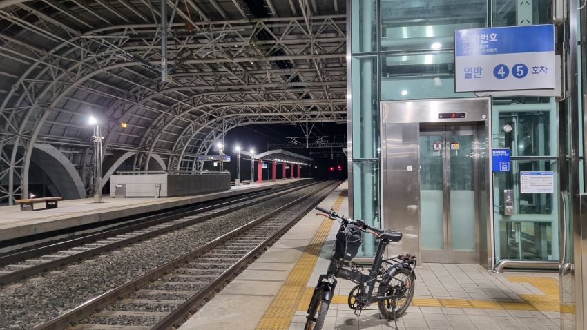 도고온천역 플랫폼, 야경, 위로 보이는 철구조물, 우측에 엘리베이터, 그 앞에 4, 5호차 승차위치 안내판, 무궁화호가 진입하고 있다는 스피커 안내방송, 승차대 노란선 뒤에 세워놓은 자전거,