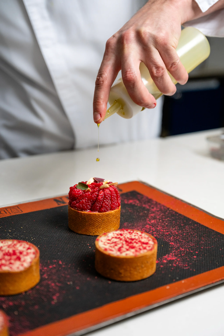 이미지 출처: AI 생성 이미지(콘텐츠 이해를 돕기 위한 시각 자료, 실제 인물을 특정하지 않은 AI 생성 이미지) 제과산업기사 실기 작업과 유사한 디저트 제작 과정으로, 타르트 위에 소스를 더해 마무리하는 제과 기술자의 섬세한 작업 장면을 보여주는 이미지입니다.