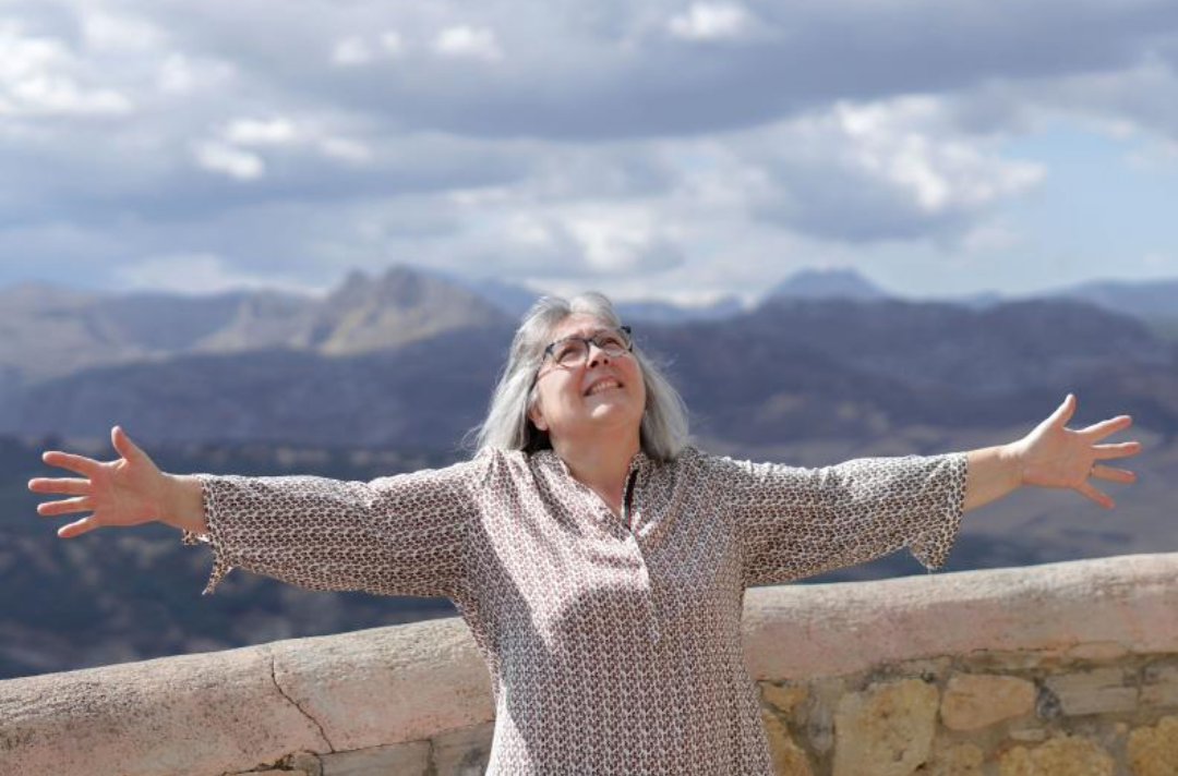 한 노인 여성이 팔을 넓게 벌리고 하늘을 바라보고 있다.