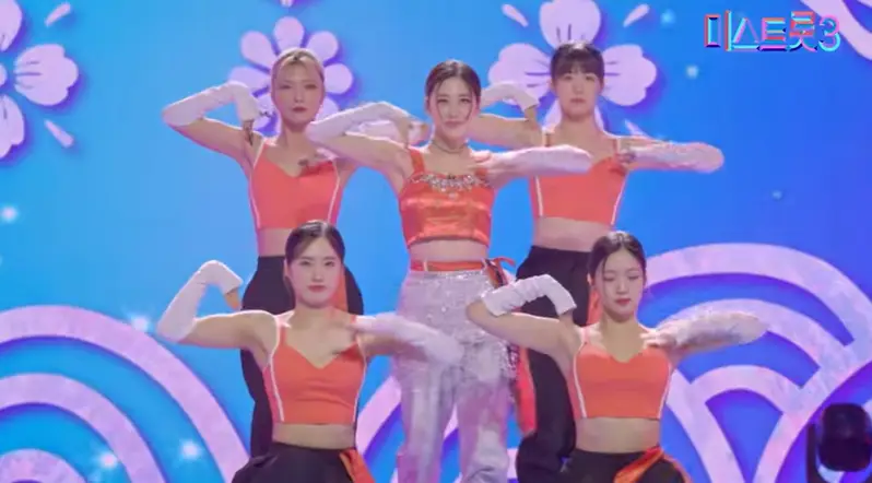 미스트롯3- 파란배경 앞 백댄서 4명 가운데 주황 나시 티에 흰바지 왼손으로 마이크를 들고 머리를 뒤로 하나로 묶은채 노래하는 윤서령