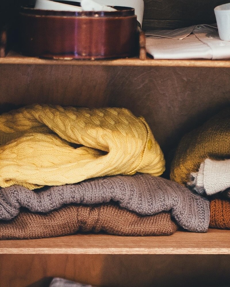 나무 선반 위에 노란색, 회색, 갈색 등 다양한 색상의 두툼한 니트와 스웨터들이 깔끔하게 접혀서 쌓여 있는 옷장 내부 모습