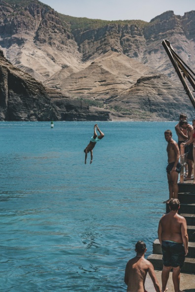 웅장한 화산암 절벽이 병풍처럼 둘러싸인 스페인 그란 카나리아의 푸른 바다에서 한 남성이 공중 다이빙을 하고 있는 모습. 오른쪽 계단에는 수영복을 입은 사람들이 다이빙을 지켜보고 있음.