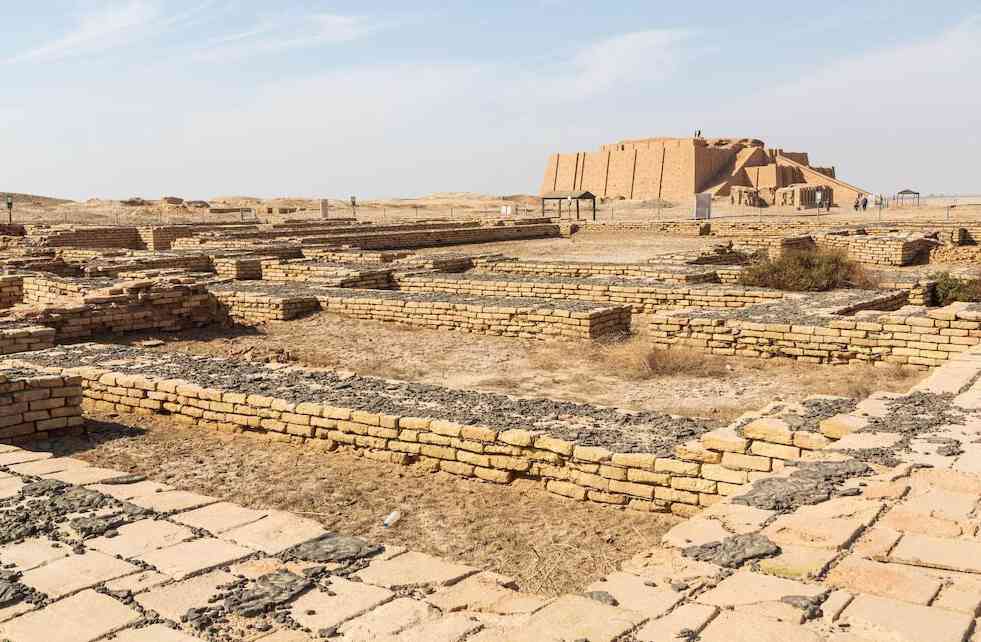 배경에 우르의 지구라트가 솟아 있고, 앞쪽에는 고대 수메르 도시의 석조 건물 기초 유적이 펼쳐진 모습. 건조한 지형과 명확한 하늘이 인상적이다