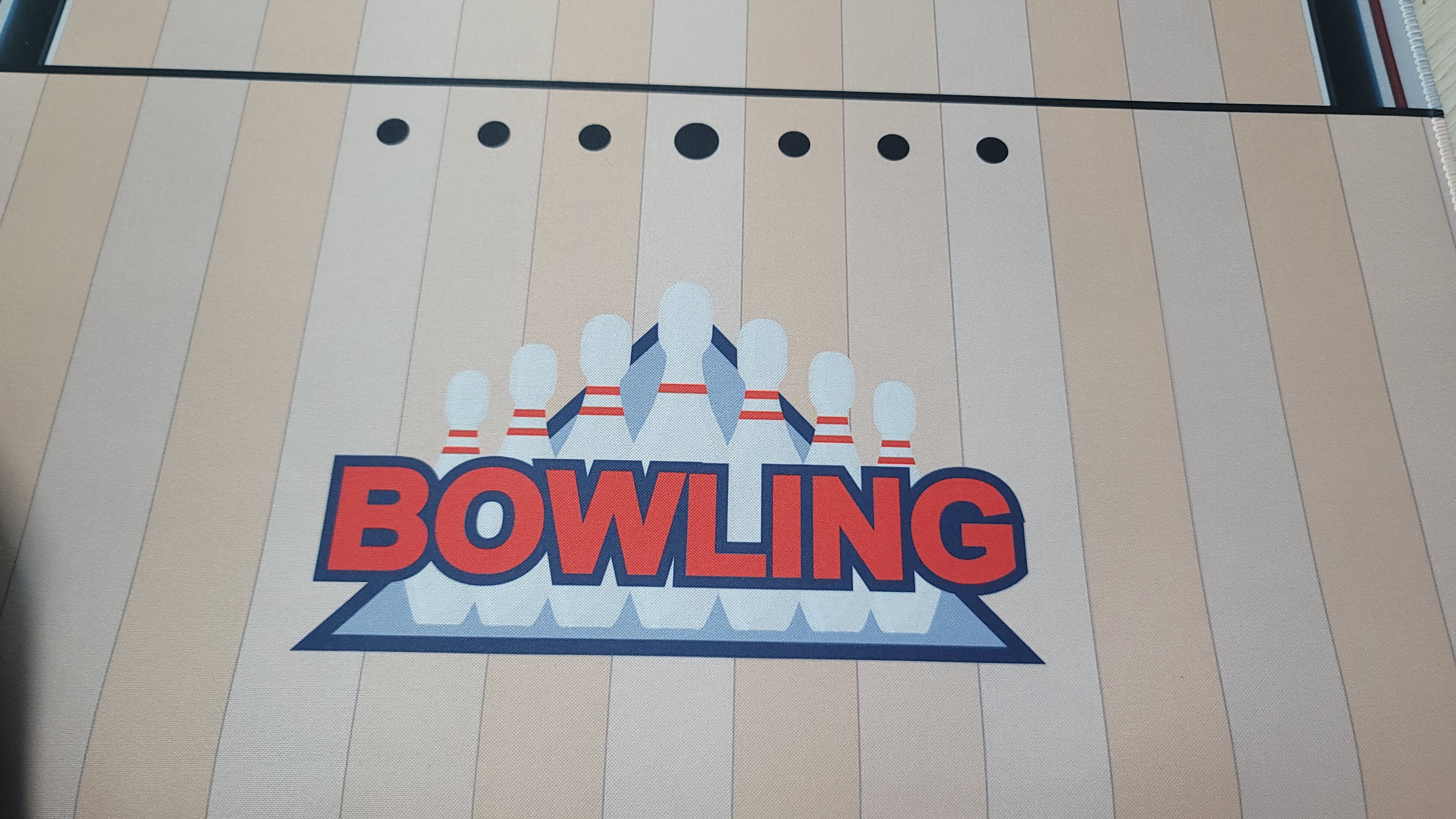 볼링 매트의 시작 부분을 근접 촬영한 사진입니다. 매트 위에는 강렬한 붉은색 글씨로 'BOWLING'이라는 영문 텍스트가 적혀 있고, 그 뒤로 볼링 핀들이 세워져 있는 그래픽이 인쇄되어 있어 실제 볼링장 레인 같은 느낌을 줍니다.