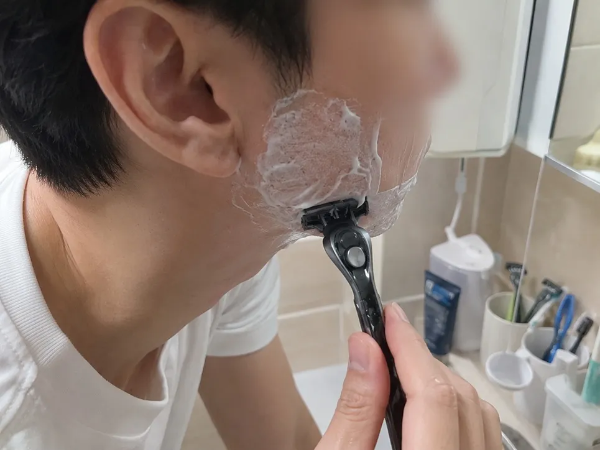하얀색 티셔츠를 입은 남성이 욕실 세면대 앞에서 볼과 턱에 하얀색 면도 폼 거품을 바르고 검은색 수동 수염 면도기를 손에 쥔 채 면도를 하고 있는 오른쪽 측면 모습 사진. 배경으로 세면대 위 컵에 꽂힌 칫솔과 면도기 등 욕실 용품들이 보이는 일상 촬영 이미지.