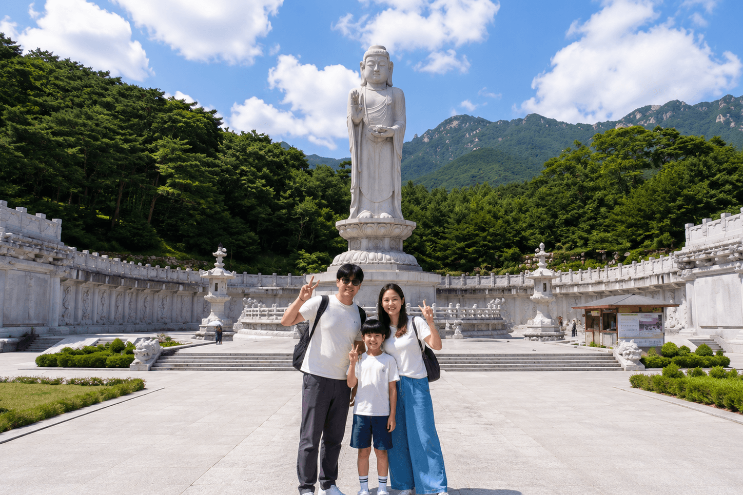 맑은 여름 하늘 아래, 대구 동화사 통일약사여래대불 앞 광장에서 하얀 티셔츠를 입은 세 가족이 브이 포즈를 하며 서 있다. 뒤에는 흰색의 대형 입상 불상과 석조 난간, 푸른 산과 숲이 보인다.