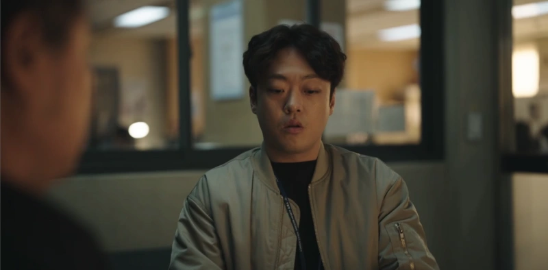 어느 회의실을 배경으로 가운데 가르마 머리에 회색 점퍼를 입고 앉아 있는 드라마 유괴의 날 채정만 캐릭터